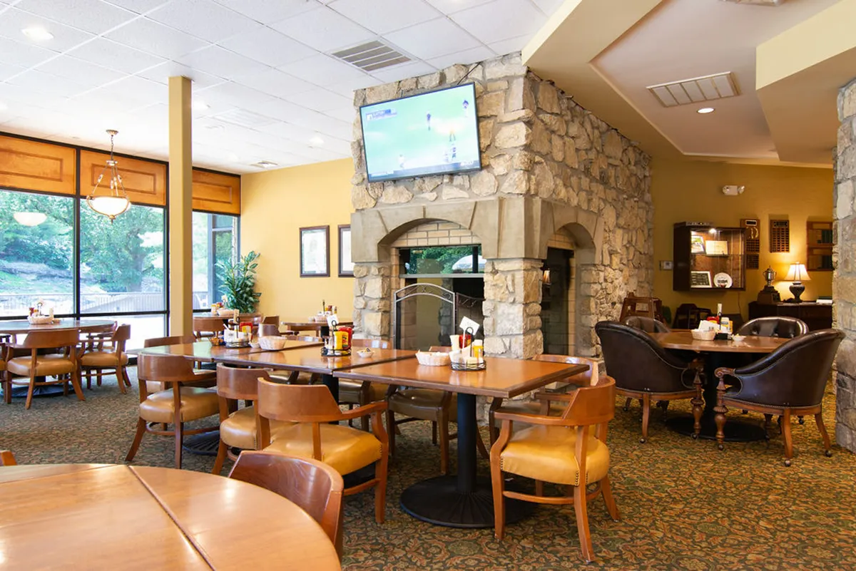 A warm interior scene inside the clubhouse featuring a stone fireplace, large windows, wooden tables, and a television for sports viewing. The communal atmosphere suggests a welcoming place for post-round meals or socializing after a day on the course.