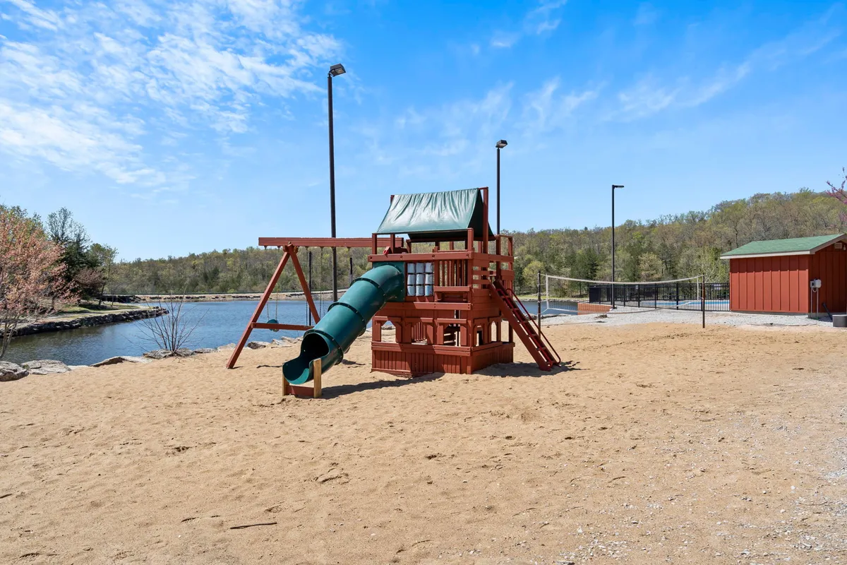 A vibrant playground with slides and climbing structures set on a sandy base, positioned beside a water body. Families can be seen enjoying the play area, and the natural backdrop signifies its integration into the greater outdoor environment. The mood is joyful, playful, and welcoming for children and families alike.