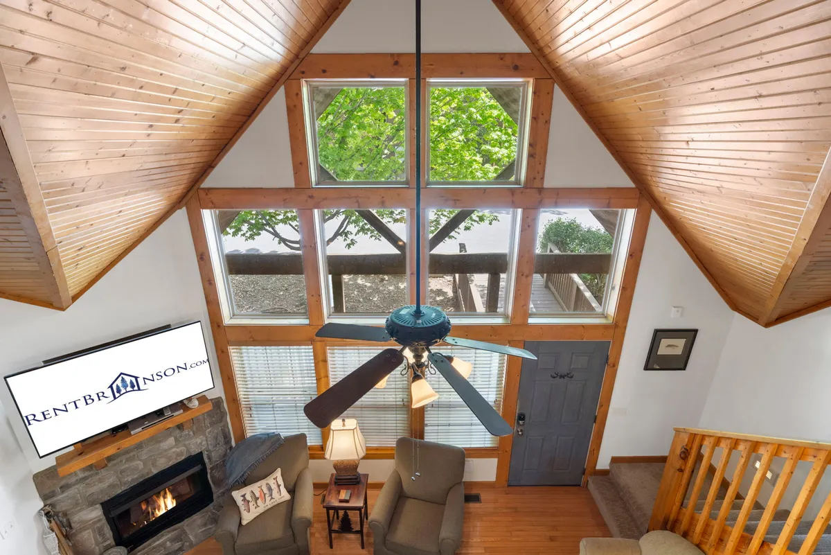 A welcoming living area with a stone fireplace, ceiling fan, large windows offering a view of nature, and inviting seating arranged for socializing and relaxation. The space feels open and perfect for both lively conversations and peaceful downtime.