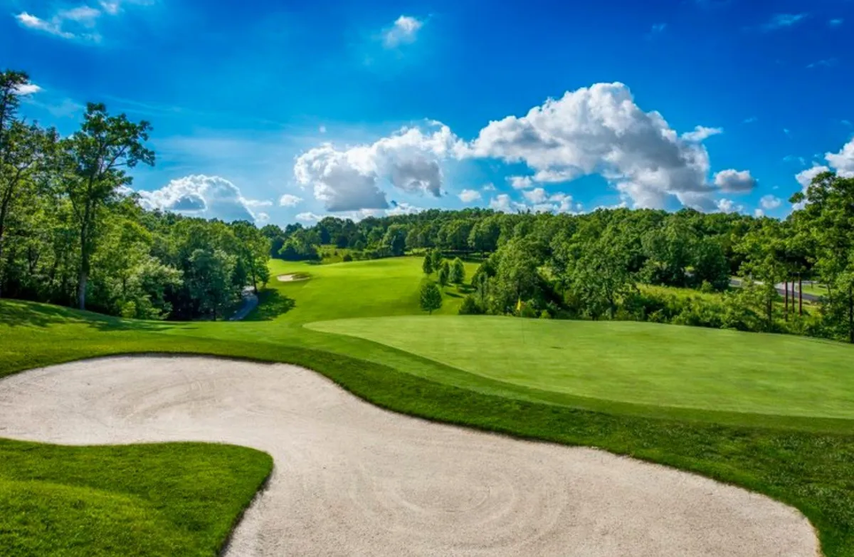 An exterior shot of a lush green fairway bordered by a sand bunker under a vibrant blue sky dotted with clouds. The open expanse of manicured grass and gentle contours suggest a well-maintained golf course, ideal for both casual and serious play.