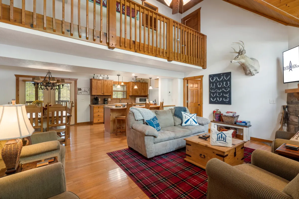 A spacious living room featuring a comfortable sofa, wooden dining table, fireplace with mounted decor, plaid rug, and large windows that flood the area with daylight. The setting is ideal for shared stories by the fire, group activities, or simply unwinding after a day out.