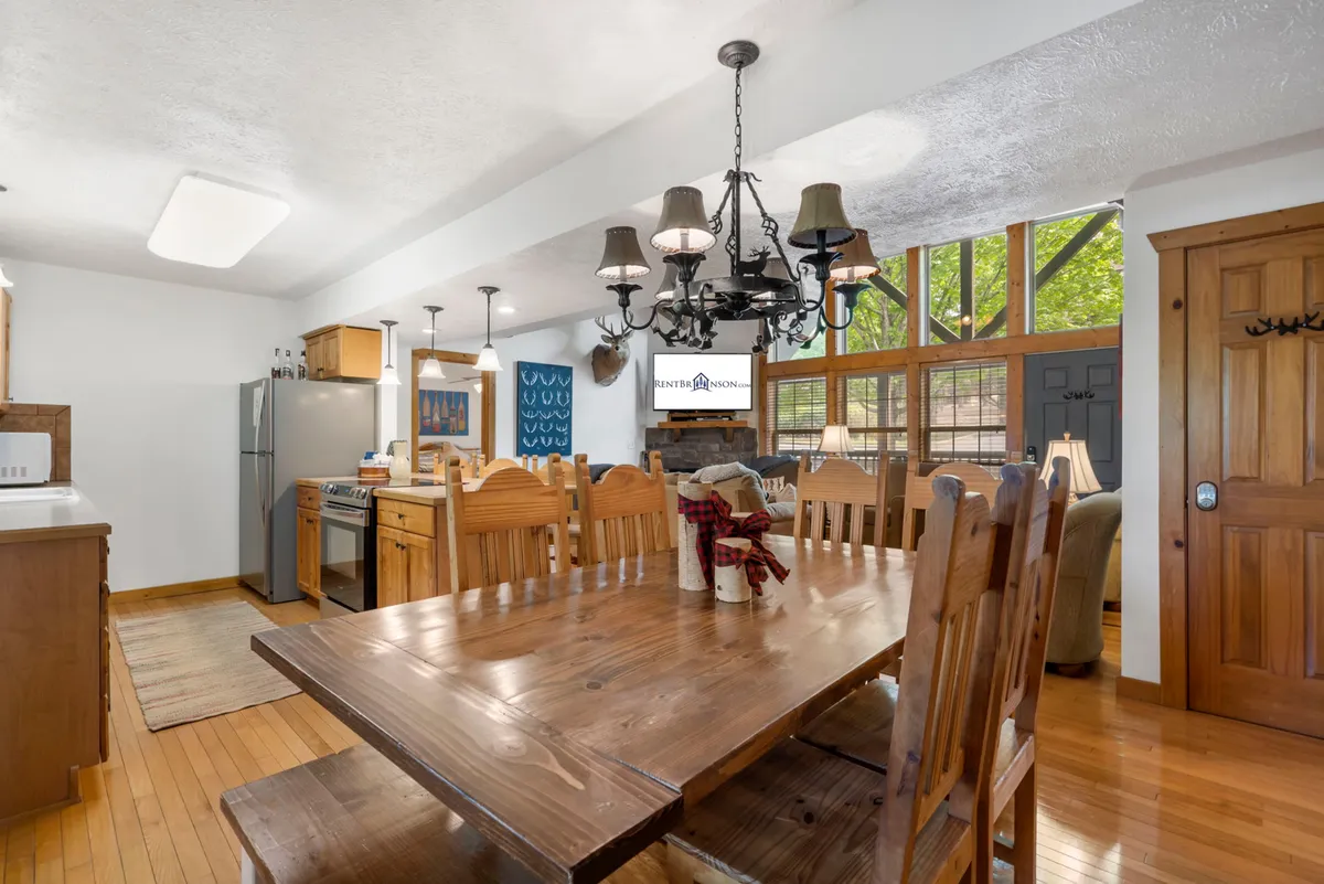 A beautifully lit open-concept dining and kitchen area featuring a large wooden dining table beneath a striking chandelier. The backdrop reveals modern kitchen appliances and expansive windows, creating a warm, communal space ideal for gatherings and shared meals.