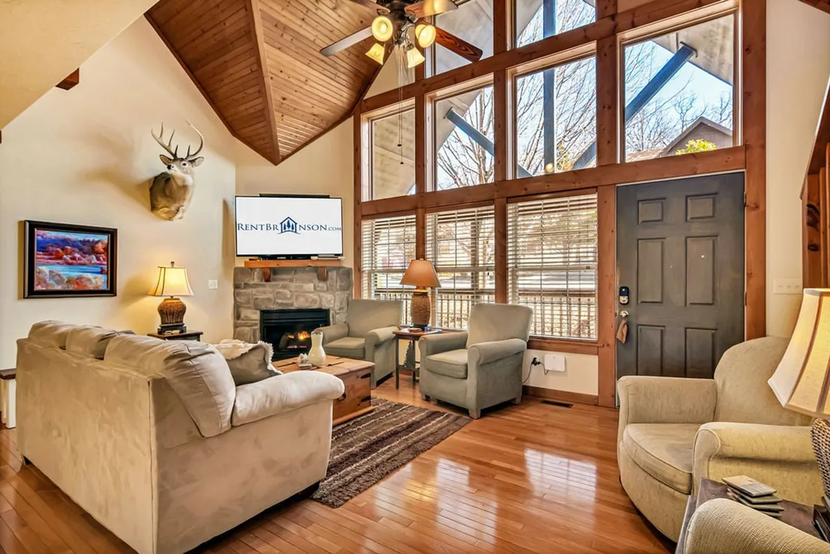 A plush living room featuring a comfortable sofa set, a glowing fireplace, mounted deer head decor, a large smart TV, and expansive windows that allow sunlight to fill the space. The arrangement encourages both group gatherings and relaxed individual moments.
