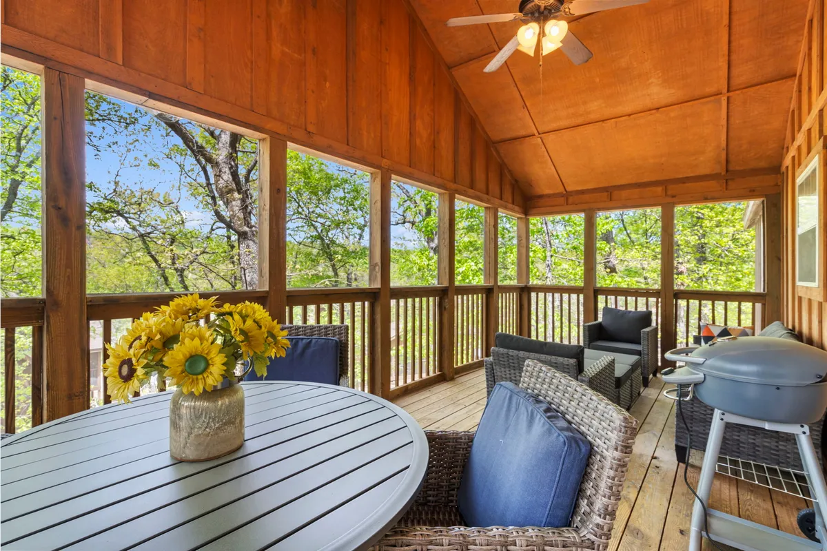 The image captures the screened-in porch of the cabin, featuring a welcoming outdoor dining table adorned with a cheerful sunflower arrangement, comfortable seating perfect for relaxing with friends and family, and a grill ready for a barbecue. Beyond the porch, lush greenery hints at the scenic natural setting, creating an inviting atmosphere for enjoying meals and nature views.