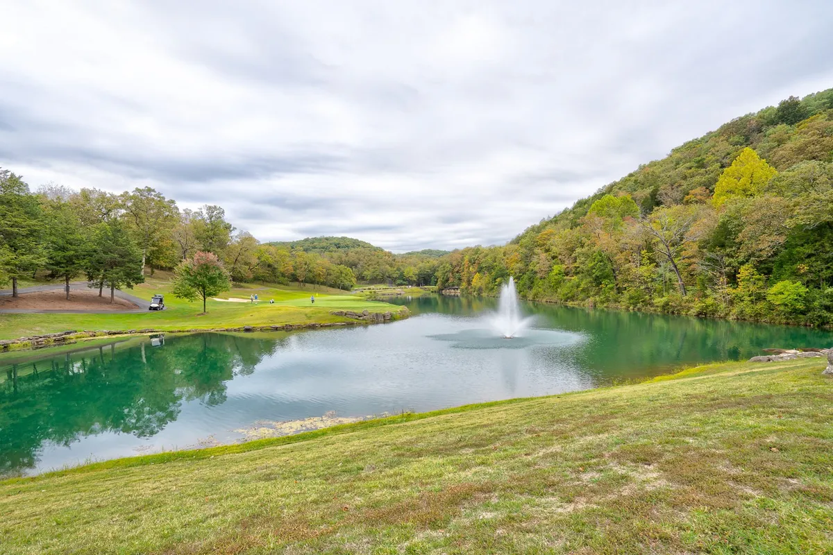 A serene lakeside view with a fountain, lush greenery, and gentle hills in the background. The image evokes peaceful outdoor recreation—fishing, walking, or simply enjoying sunrise or sunset by the water.