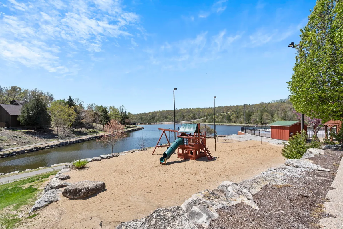 An inviting, playful scene with playground equipment set on a sand area beside a tranquil water body, surrounded by greenery. Families and children are encouraged to play, picnic, and relax by the water’s edge.