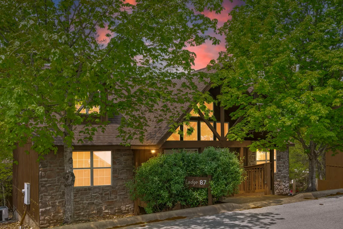 A welcoming lodge exterior with warm wooden architecture, surrounded by lush greenery and warmly lit windows. The inviting porch suggests a perfect place to relax after days filled with Branson’s adventures, highlighting the home’s cozy retreat vibe.